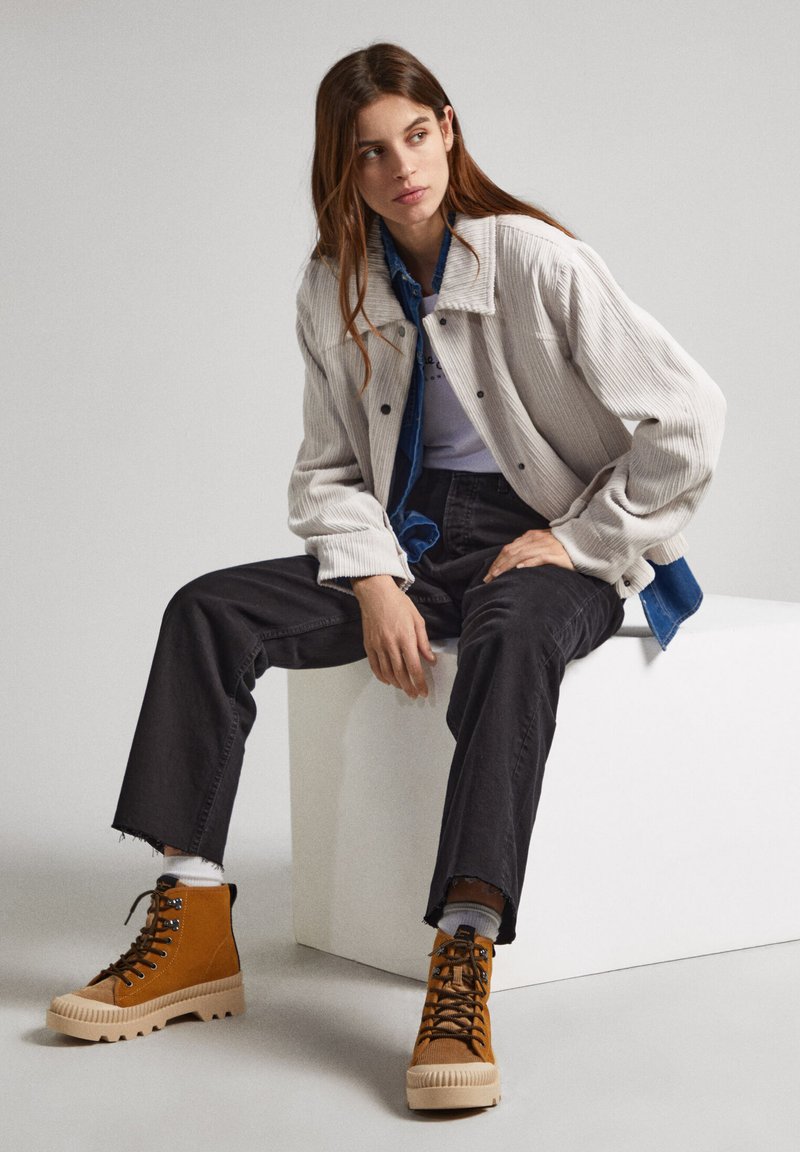 Chaqueta de pana beige sobre una camisa azul, jeans negros con dobladillos deshilachados y botas altas marrones con suelas crema. Fondo neutro.