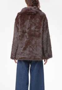 Cappotto in pelliccia sintetica marrone con colletto, maniche lunghe e una texture shaggy. Abbinato a pantaloni in denim a gamba larga.