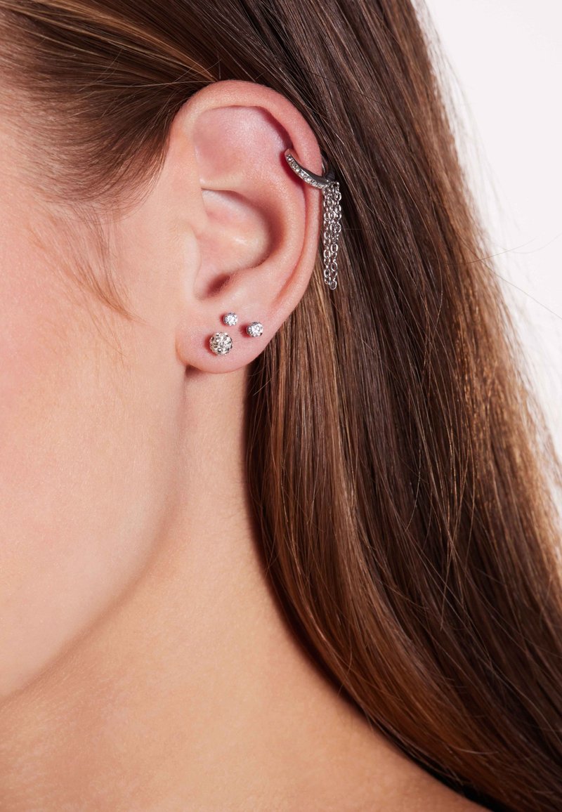 Zilveren oorcuff met een ketting, trio van kleine stud oorbellen: één bolvormig, één met een kristal en één vlak, allemaal met een glanzende afwerking.