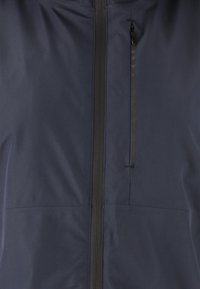 Donkerblauwe jas met een rits aan de voorkant, inclusief een klein ritszakje op de borst en gemaakt van glad, lichtgewicht materiaal met een subtiele textuur.