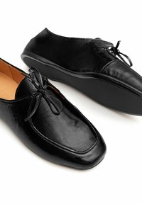 Scarpe in pelle nera con una finitura liscia, punta tonda e dettaglio a fiocco frontale. La suola è piatta e leggermente rialzata al tallone.
