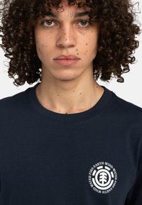 Marineblaue Baumwoll-T-Shirt mit rundem Halsausschnitt, ausgestattet mit einem zentrierten weißen, runden Logo mit Text und einer Baumgrafik auf der linken Brust.
