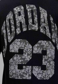 Juodas marškinėliai su baltu nėriniuotu raštu ant skaičių "23" ir "JORDAN" drąsia sportine šriftu.