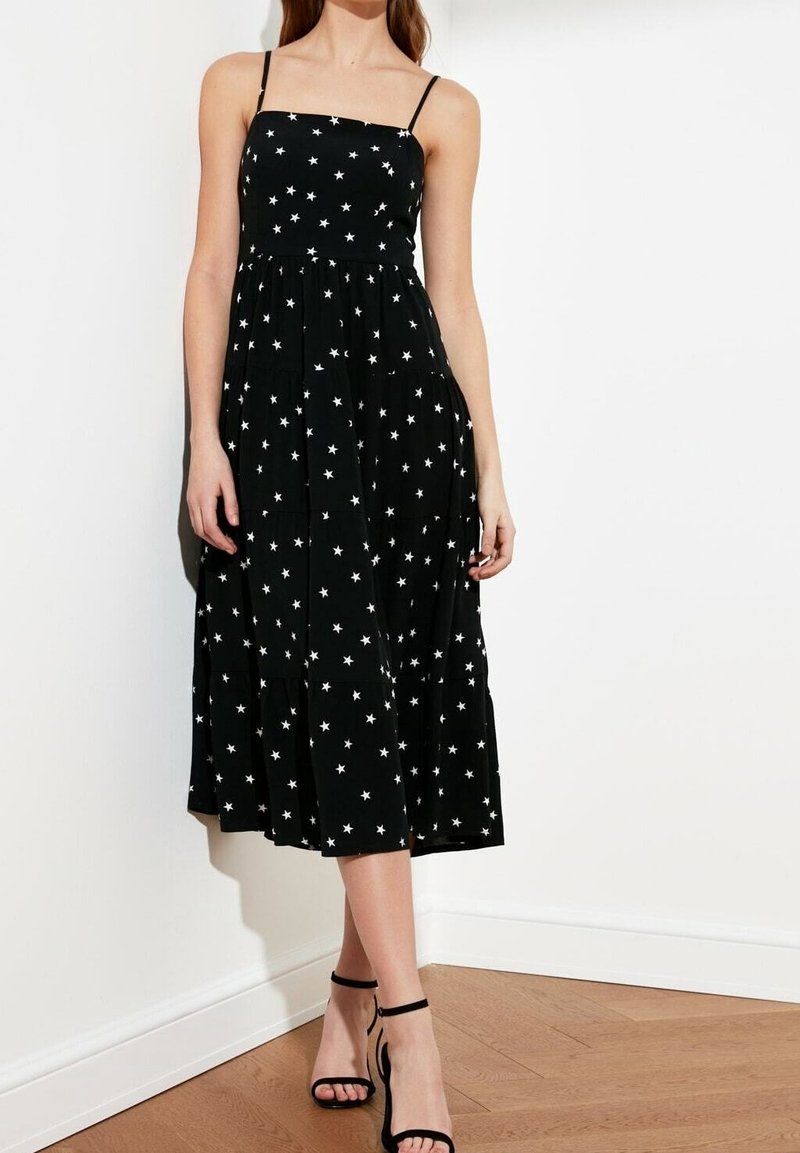 Kobieta ubrana w czarną midi sukienkę w białe gwiazdki i cienkie ramiączka, w połączeniu z czarnymi sandałami na wysokim obcasie, stoi w pomieszczeniu.