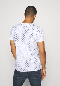 Hombre visto de espaldas con una camiseta de manga corta blanca con un pequeño patrón de banderas azules y jeans azul oscuro.