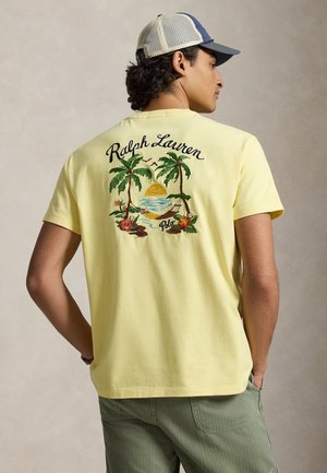 Άνδρας με κίτρινη μπλούζα με κεντητό τροπικό ηλιοβασίλεμα, φοίνικες, αιώρα και την επιγραφή "Ralph Lauren Polo" στην πλάτη, μπλε καπέλο.