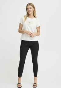 Biely tričko s jemným kvetinovým vzorom, skombinované s čiernymi úzkymi nohavicami. Modelka stojí so zlomenými rukami, má na sebe čierne sandále na opätku.
