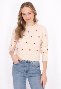Maglione color crema con cuori ricamati rossi, scollatura rotonda e polsini a coste, abbinato a jeans in denim a vita alta.