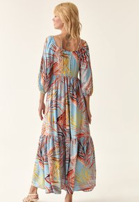 Maxikleid aus hellblauem Stoff mit auffälligen Mustern von tropischen Blättern in Rot, Orange und Gelb, puffärmelig und mit einem gestuften Saum.