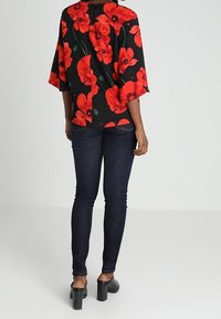 Blouse noire avec un grand motif floral rouge, manches chauve-souris amples et fermeture dans le dos. Associée à un jean skinny foncé et des talons chunky noirs.