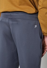Pantaloni sportivi grigi in tessuto liscio, con vita elastica e un logo discreto sulla coscia sinistra.