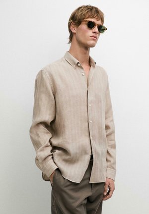 Junger Mann in beigem Hemd mit Knöpfen, brauner Hose und Sonnenbrille, stehend mit einer Hand in der Tasche vor einfachem Hintergrund.