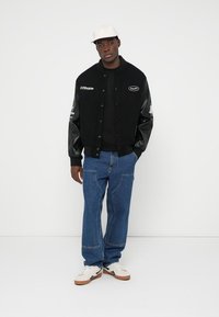 Fekete sportos kabát bőr ujjal, hímzett logókkal, fekete kereknyakú póló, kék farmernadrág és fehér sportcipő. A modellen fehér sapka van.