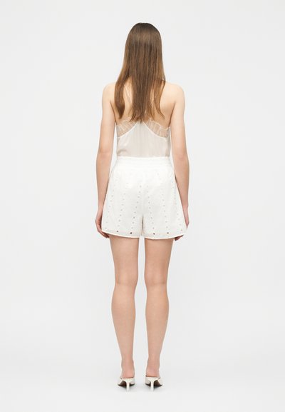 Kobieta z długimi brązowymi włosami, ubrana w bezrękawnik w kolorze beżowym z koronkowymi zdobieniami oraz białe szorty z wysokim stanem, stoi tyłem.