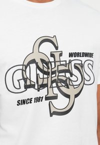 Hvid T-shirt med lagdelte "GUESS" bogstaver, store beige sammenkoblede G'er, samt sort tekst "WORLDWIDE" og "SINCE 1981" på brystet.