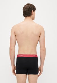 Mustat bokserialushousut, joissa on punainen vyötärönauha ja "Calvin Klein" -kirjainlogo. Sileä pinta, vartalonmyötäinen malli ja reisien puoliväliin ulottuva pituus.