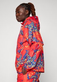Chaqueta roja con patrones abstractos en azul y amarillo, con capucha ajustable, puños elásticos y bolsillos laterales. Material ligero.