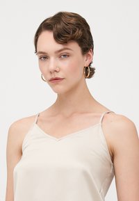 Beige satengamisole med tynne stropper, v-hals, glattet tekstur og minimal sømdetalj. Gull hoop øredobber brukt som aksent.