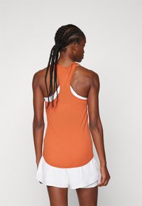 Orange atletisk tanktop med racerback design og hvide detaljer, parret med hvide shorts. Glat tekstur, tætsiddende facon og buet kant.