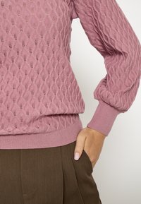 Vrouw draagt een roze gebreide trui met textuur en pofmouwen, gecombineerd met bruine geplooide broek, hand rustend in de zak met lichtroze nagels.