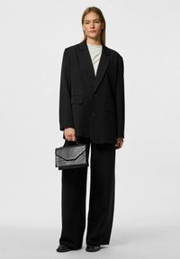 Blazer nero oversize con bottoni frontali e due tasche a toppa, abbinato a pantaloni neri a gamba larga e una borsa nera testurizzata.