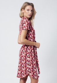 Abito floreale rosso con maniche corte e design avvolgente. Presenta fiori rosa e bianchi su uno sfondo vivace, insieme a un dettaglio con spacco laterale.