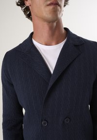 Marineblauwe gebreide jas met een gestructureerd visgraatpatroon, dubbele sluiting, reverskraag en twee knopen aan de voorkant.