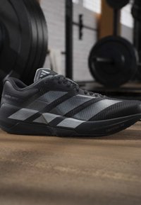 Μαύρο και γκρι παπούτσι Adidas για τρέξιμο με σόλα Lightstrike Pro, τοποθετημένο στο πάτωμα γυμναστηρίου με εξοπλισμό άρσης βαρών θολωμένο στο φόντο.
