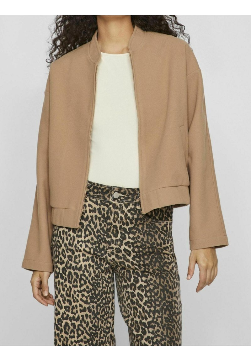 Femme portant une veste beige zippée sur un top blanc, associée à un pantalon taille haute imprimé léopard, debout, les bras détendus.