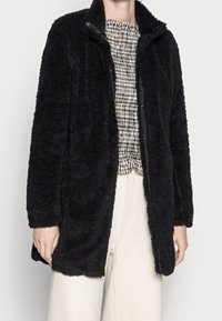 Schwarzer, flauschiger Mantel mit langen Linien und weicher Textur, mit Knopfverschluss und Kragen, getragen über einem gemusterten Oberteil und hellen Hosen.