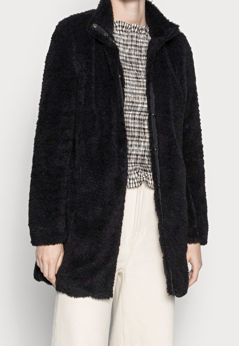 Schwarzer, flauschiger Mantel mit langen Linien und weicher Textur, mit Knopfverschluss und Kragen, getragen über einem gemusterten Oberteil und hellen Hosen.