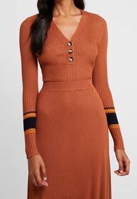 Dlouhé rukávy, žebrované šaty v terakotové barvě s výstřihem do V, třemi dekoračními knoflíky a pruhovanými akcenty v námořnické, žluté a oranžové barvě na rukávech.