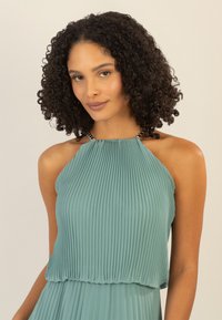 Ein ärmelloses, plissiertes Kleid in Türkis mit hohem Ausschnitt, Kettenakzenten an den Schultern und fließendem Stoff, der Bewegung erzeugt.