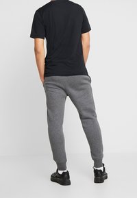 Mann in einem schwarzen T-Shirt, grauen Jogginghosen, weißen Socken und schwarzen Turnschuhen, der mit den Händen in den Taschen abgewandt steht.
