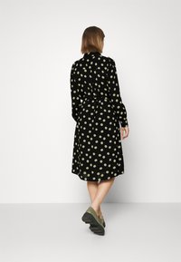 Abito nero con pois verdi, maniche lunghe, vita con fiocco e orlo fino al ginocchio. Il tessuto appare liscio e leggero.