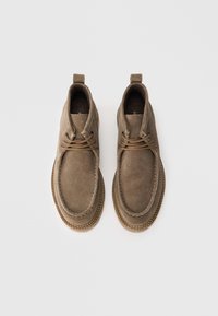 Et par lysebrune suede ankelstøvler med runde tåspidser, snørebånd foran og brune syede gummisåler på en hvid baggrund.