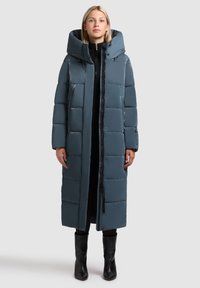 Pikk, pehmealt soojustatud mantel tuhmises sinises värvitoonis, kõrge kraega ja suure kapuutsiga. Omaduseks on lukuga sulgemine ja küljetaskud. Sile, läikiv tekstuur.