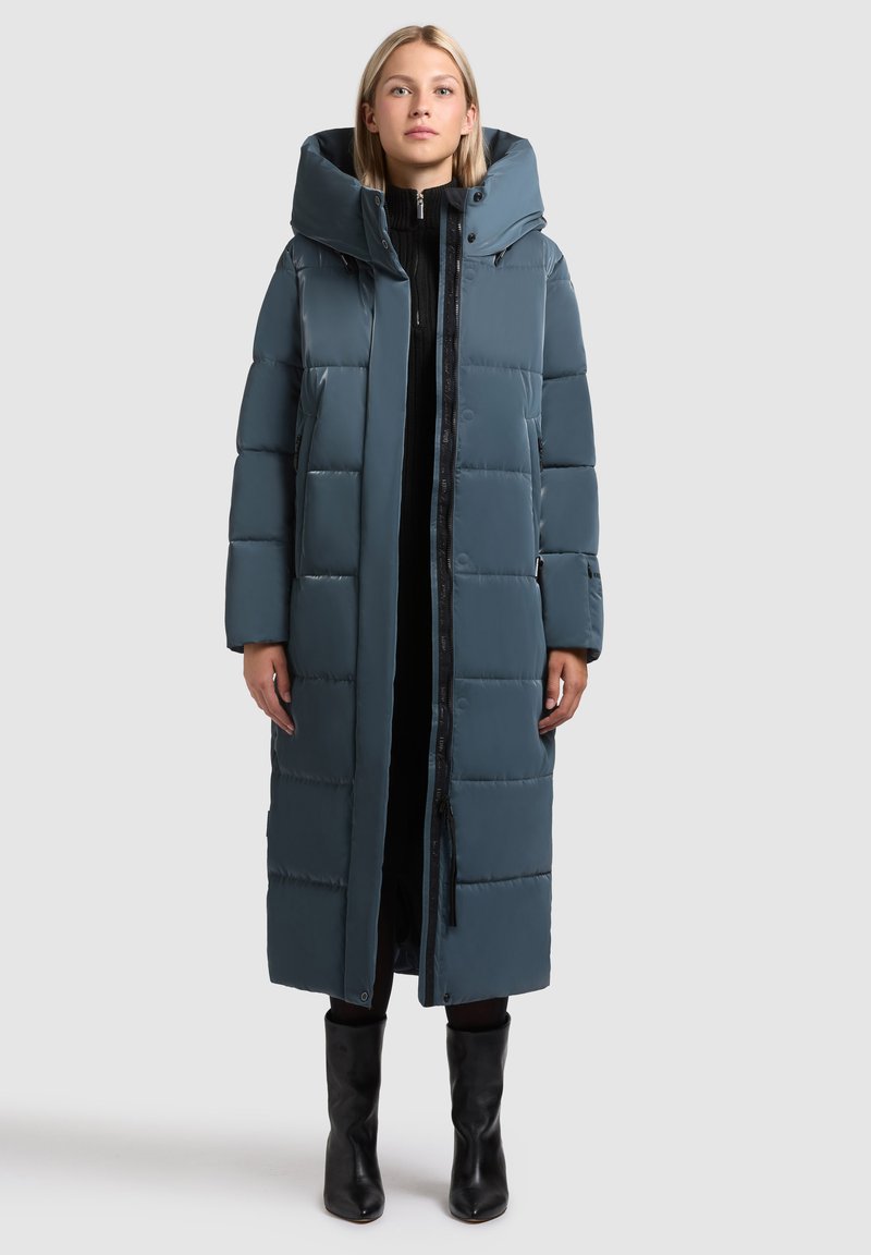 Pikk, pehmealt soojustatud mantel tuhmises sinises värvitoonis, kõrge kraega ja suure kapuutsiga. Omaduseks on lukuga sulgemine ja küljetaskud. Sile, läikiv tekstuur.