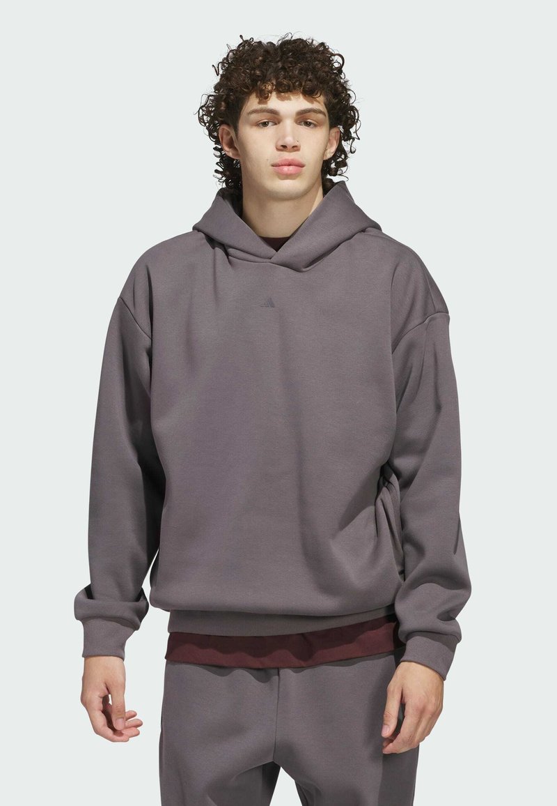 Siva dukserica od mekog materijala, s opuštenim krojem, kapuljačom s vezicom i diskretnim logo detaljem. Nošena preko vino crvene majice.