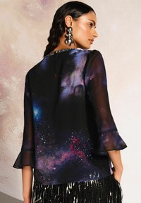 Blusa nera trasparente con stampa galaxia, con maniche a campana e dettaglio a occhiello sul retro. Accentuata da un colletto gioiello.