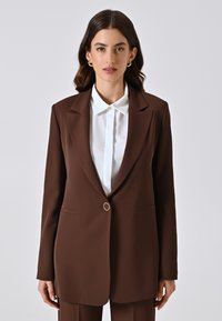 Blazer marrone con un singolo bottone dorato, rever a scollo a V, due tasche frontali e vestibilità su misura sopra una camicia bianca con bottoni.