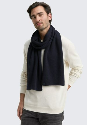 Navy ribbed knit scarf draped around the neck, made from soft material, paired with a light-colored sweater and grey jeans.