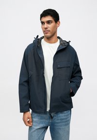 Granatowa kurtka wiatrówka z zamkiem z przodu, kieszenią na piersi i kratowanym podszyciem kaptura, wykonana z lekkiego, gładkiego materiału.