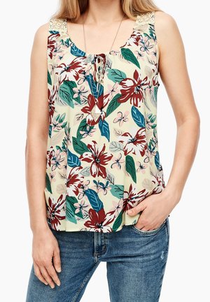 Kvinde iført ærmeløs blomsterprintet top med blonde stropper og blå jeans, den ene hånd i lommen, og lagdelte geometriske vedhæng halskæder.
