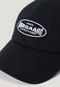 Căciulă de baseball neagră din bumbac, cu vizieră curbată. Prezintă un logo brodat alb: „MADS NORGAAARD COPENHAGEN” pe partea din față.
