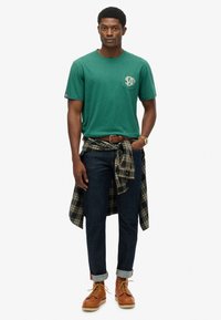 Camiseta verde de manga corta con detalle de bolsillo, jeans oscuros, camisa de cuadros atada a la cintura y botas marrones con cordones.