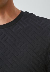 Sort kortærmet T-shirt med et struktureret geometrisk mønster, der har en ribbet halsudskæring og glat stof. Nærbillede af materialet.