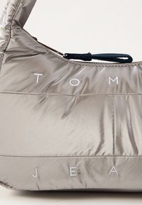 Sudrabota pūkains rokassomu ar spīdīgu apdari, kurai ir balti izšūti burti "TOM JEAN" un augšējais rokturis ar tumši zilām akcentiem.