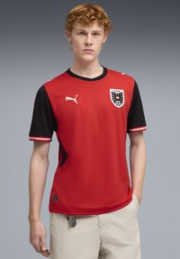 Κόκκινη και μαύρη αθλητική φανέλα με κοντό μανίκι, με στρογγυλή λαιμόκοψη, πλαϊνές λεπτομέρειες και έμβλημα στο αριστερό στήθος. Κατασκευασμένη από διαπνέον υλικό.