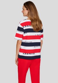 Gestreiftes T-Shirt in Rot, Weiß und Marineblau mit grafischem Text; kurze Ärmel und lockere Passform. Kombiniert mit roten Hosen.